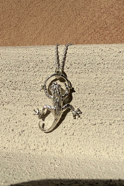 Silver gecko pendant on a chain against a beige wall