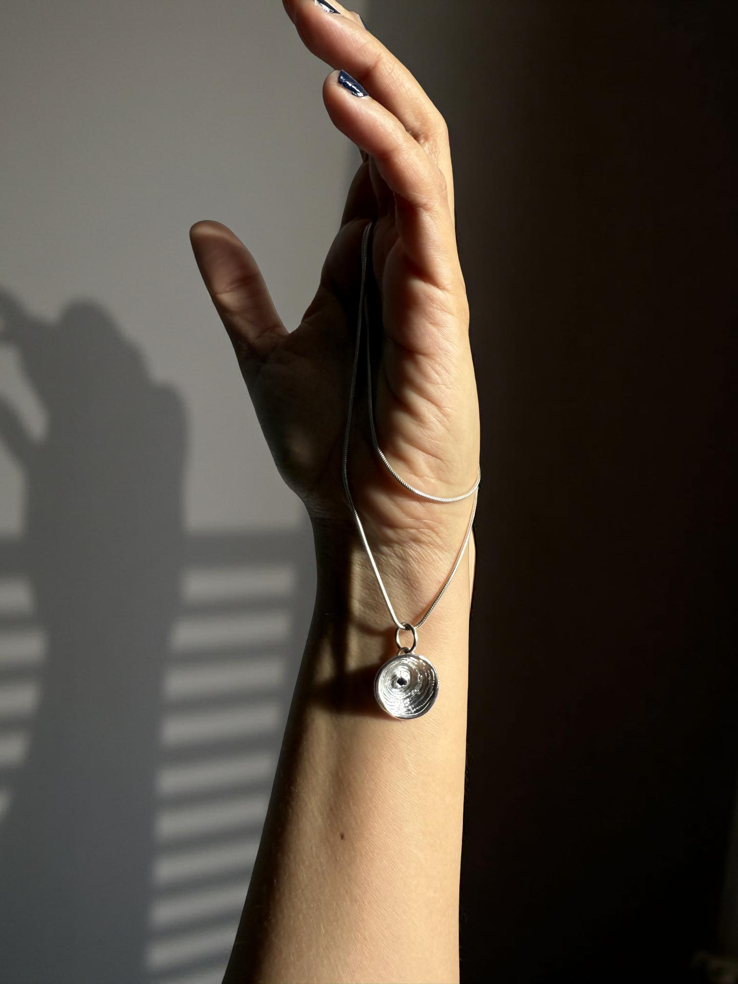 Hand wearing a silver necklace with a pendant against a gray background