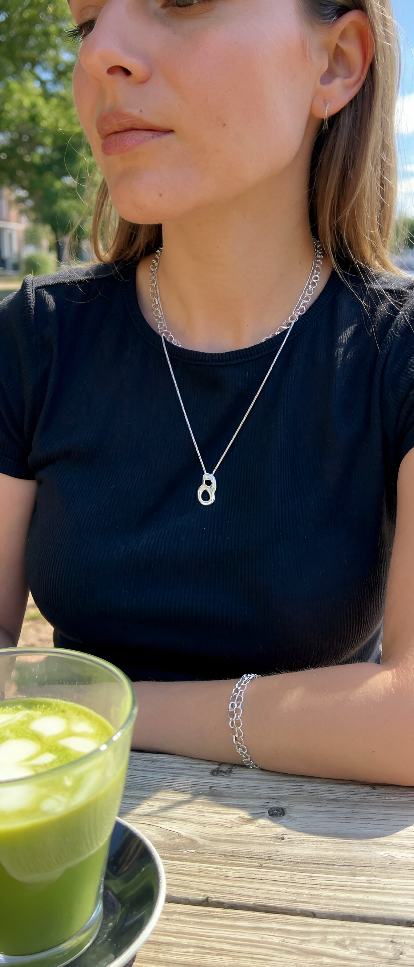 Woman sitting at a table with a green drink, wearing a black top and silver necklace.