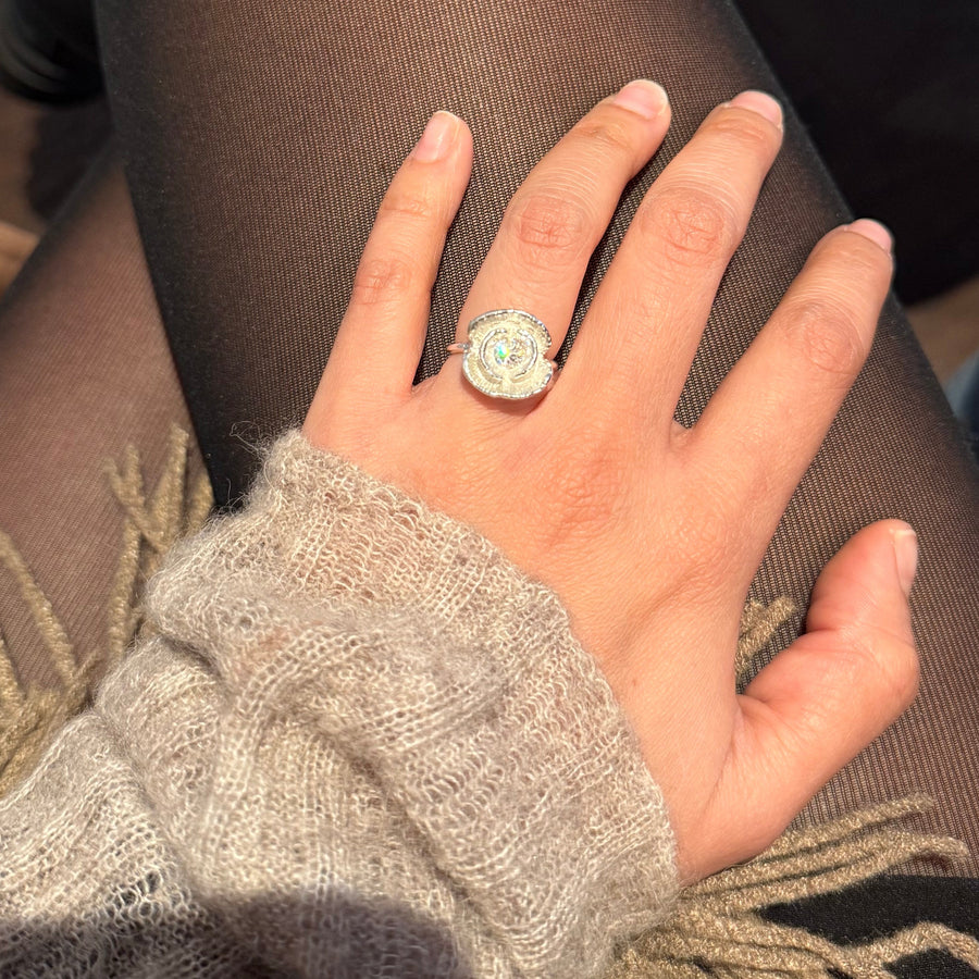 A close-up shot of a person's hand resting on their thigh, wearing a large silver-toned ring with a round clear gemstone. The person is wearing black tights, a beige knit sweater, and a brown scarf in an indoor setting.