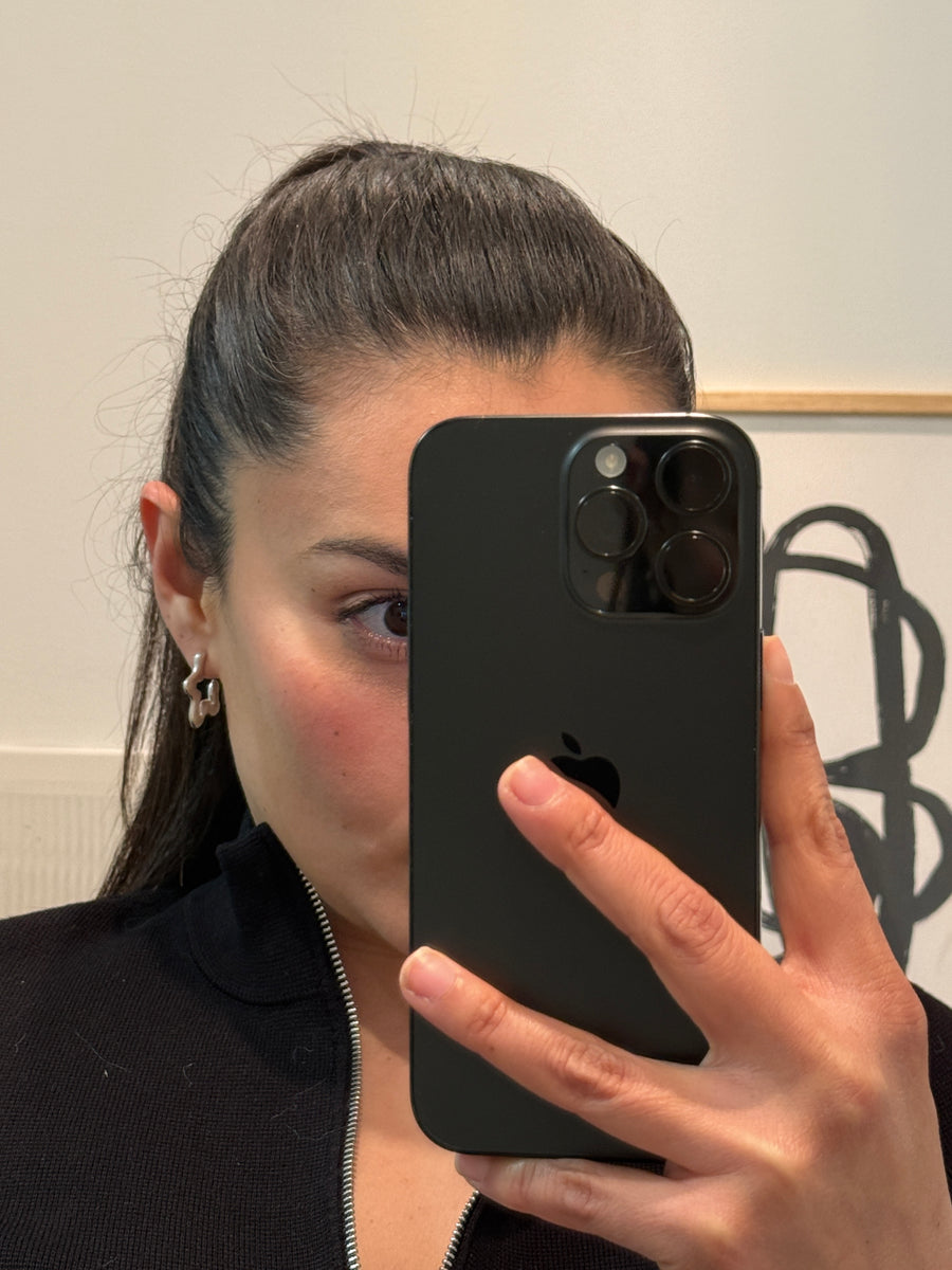 A person taking a mirror selfie with a black smartphone in front of their face. The phone has a large triple-camera array on the back and a visible Apple logo, indicating the brand is Apple.