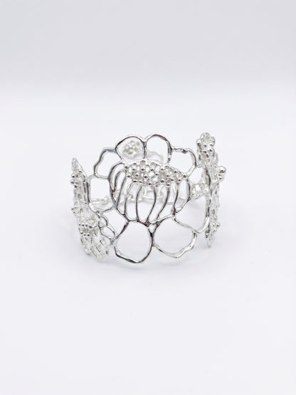 A silver-toned openwork cuff bracelet with a floral filigree-style design inspired by the floral motifs in lace, photographed on a seamless light gray background.
