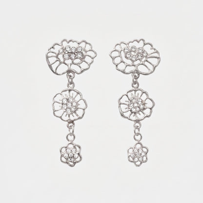 A pair of silver-toned dangle earrings with moissanites at the center, featuring three distinct floral designs inspired by lace motifs, connected by small chains, shown against a plain light gray background. 