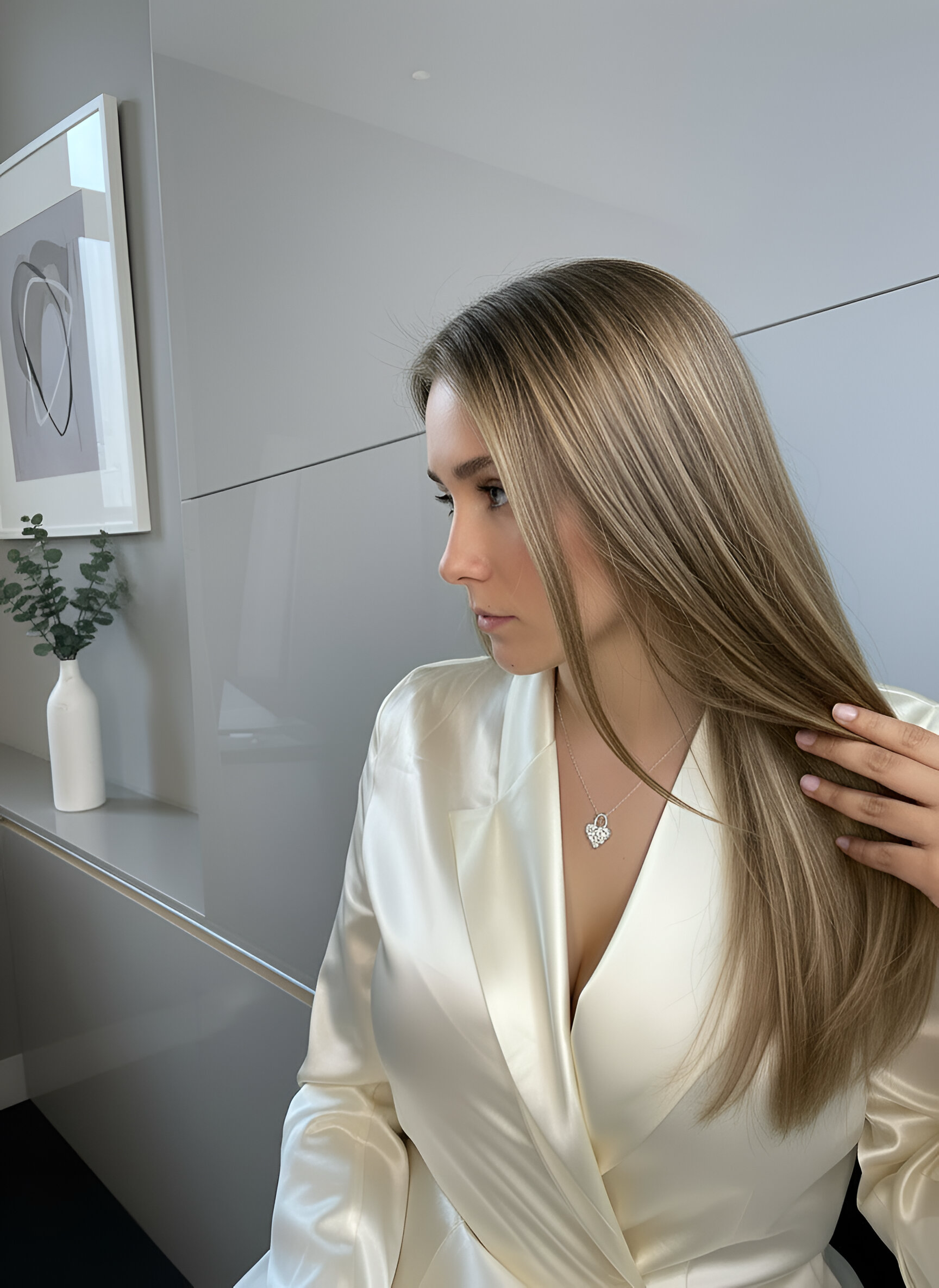 A woman in a white satin dress sitting indoors. She is wearing a heart-shaped lock necklace, cushioned heart with 7 white moissanites at each intersection.