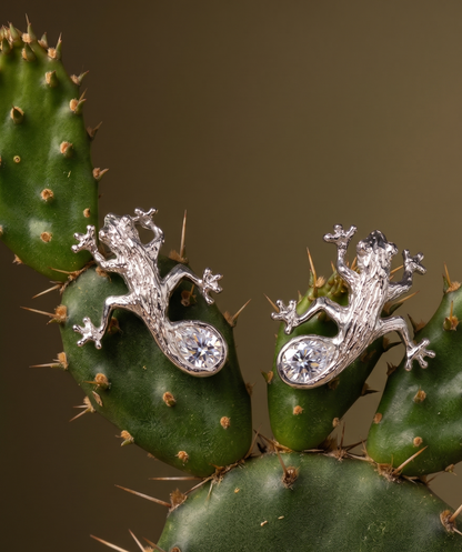 Luxury Solid sterling silver gecko-shaped earrings with a pear-shaped white moissanite stone. The earring is crafted by hand and showcased on a cactus plant.