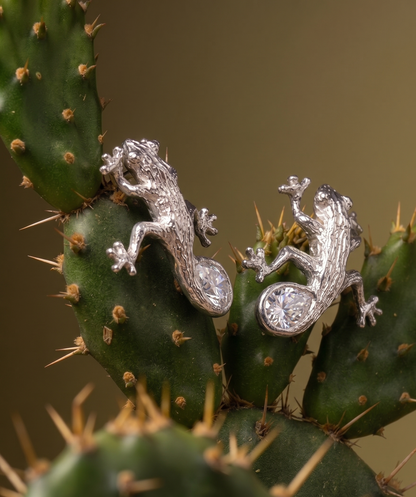 Luxury Solid sterling silver gecko-shaped earrings with a pear-shaped white moissanite stone. The earring is crafted by hand and showcased on a cactus plant.