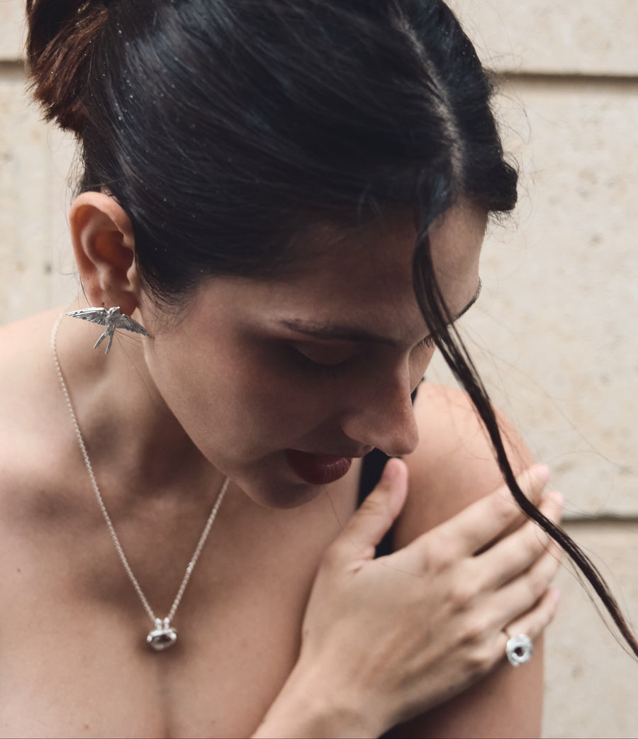 A woman with dark hair in a messy bun wearing a silver swallow earring and a poppy flower pendant necklace and matching poppy flower ring, posing against a beige stone wall in Paris.