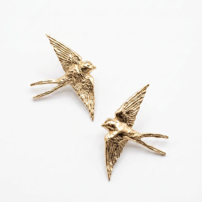 A pair of gold-toned metal earrings shaped like flying swallows, displayed on a clean white background.