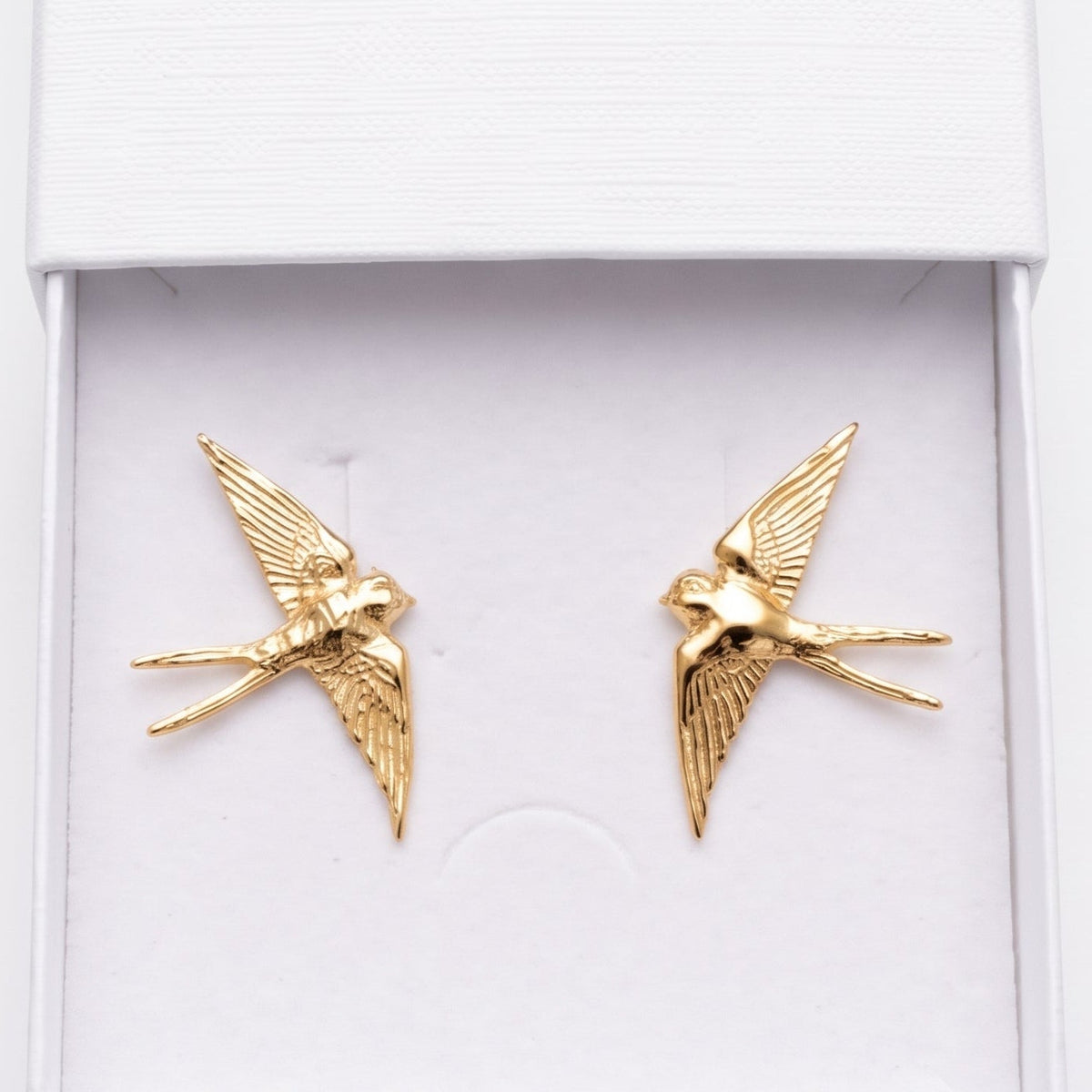 A pair of gold swallow-shaped stud earrings displayed inside an open white jewelry box. The embossed brand name 'nounou' is visible on the inside of the lid, and the box is photographed against a clean white background.