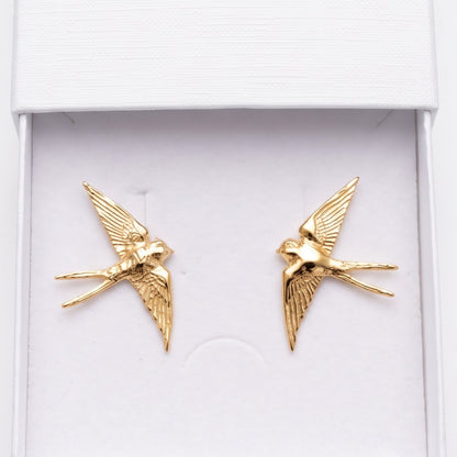 A pair of gold swallow-shaped stud earrings displayed inside an open white jewelry box. The embossed brand name 'nounou' is visible on the inside of the lid, and the box is photographed against a clean white background.
