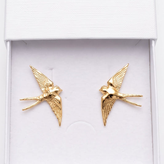 A pair of gold swallow-shaped stud earrings displayed inside an open white jewelry box. The embossed brand name 'nounou' is visible on the inside of the lid, and the box is photographed against a clean white background.