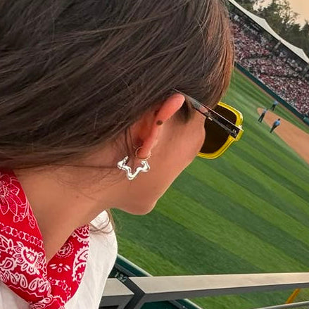 A man and woman wearing star-shaped sterling silver earrings at a baseball game, viewed from behind the man. He is wearing a bright red baseball cap with a white script logo, while she wears a red bandana and white shirt.